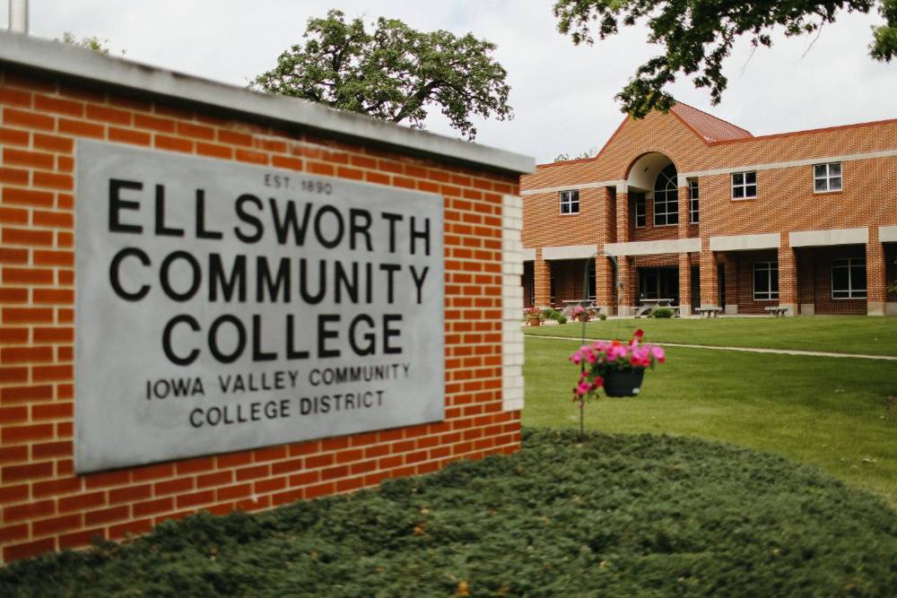 Brick monument sign reading “Ellsworth Community College” in the foreground, with a red-brick campus building, green lawn, trees, and hanging flower baskets visible in the background on an overcast day.